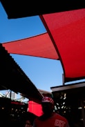 A vivid red fabric canopy contrasts against a bright blue sky. The street scene features shaded areas and silhouettes of people, with one person wearing a red and white patterned cap and a shirt with 'EST' printed on it. The surrounding structures create geometric patterns of light and shadow.
