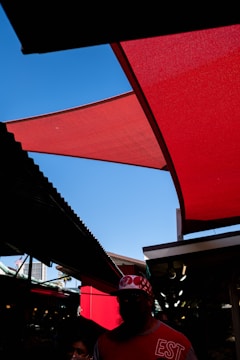 A vivid red fabric canopy contrasts against a bright blue sky. The street scene features shaded areas and silhouettes of people, with one person wearing a red and white patterned cap and a shirt with 'EST' printed on it. The surrounding structures create geometric patterns of light and shadow.