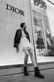 A person in a white dress and black leather jacket poses in front of a store window. The window displays two dresses on mannequins and reflects a cityscape. The word 'DIOR' is prominently featured on the wall.