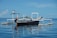 A small fishing boat is floating on calm blue waters under a clear sky. The boat is equipped with outriggers, a motor at the back, and a light fixture mounted on a pole. The water reflects the boat and sky, creating a serene and peaceful scene.