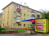 A four-story residential building with a yellow brick facade and multiple small balconies is situated in an urban area. In front of the building, there is a small street market stall with a blue and red canopy. The stall displays various fruits and vegetables, with a colorful advertisement on its side featuring images of fruits and vegetables and labeled in Cyrillic script.