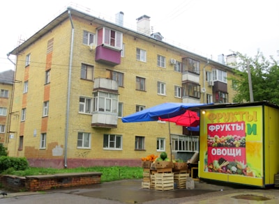 A four-story residential building with a yellow brick facade and multiple small balconies is situated in an urban area. In front of the building, there is a small street market stall with a blue and red canopy. The stall displays various fruits and vegetables, with a colorful advertisement on its side featuring images of fruits and vegetables and labeled in Cyrillic script.