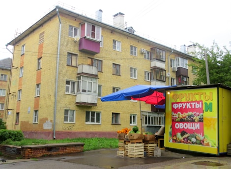 A four-story residential building with a yellow brick facade and multiple small balconies is situated in an urban area. In front of the building, there is a small street market stall with a blue and red canopy. The stall displays various fruits and vegetables, with a colorful advertisement on its side featuring images of fruits and vegetables and labeled in Cyrillic script.