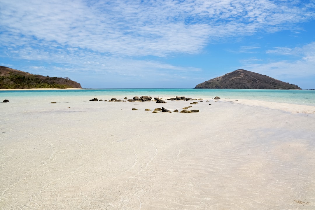 white sand beach with brown mountain in distance under blue sky and white clouds during daytime