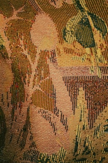 An intricately woven tapestry showcasing an abstract pattern with various shapes that resemble leaves and foliage. The texture is rich and detailed, with visible individual threads creating a sense of depth and complexity.