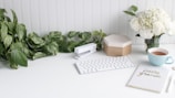A clean and organized workspace features a white keyboard, a white stapler, and a pen on a white desk. On the left, there is a lush green potted plant, while on the right, a bouquet of white roses and hydrangeas sits in a vase next to a blue teacup filled with tea. A notebook with a polka dot cover and the words 'Let's do this' written on it is also present.