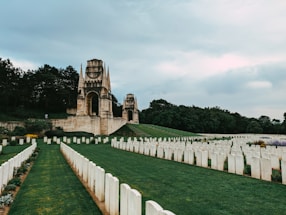 A solemn cemetery with rows of white tombstones neatly arranged on green grass. In the background, two large stone monuments with spires and arches are visible, surrounded by dense trees under a cloudy sky.