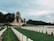 A solemn cemetery with rows of white tombstones neatly arranged on green grass. In the background, two large stone monuments with spires and arches are visible, surrounded by dense trees under a cloudy sky.
