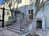 A set of brick stairs with black metal railings leads up to a door on the side of a light-colored building. The area is enclosed by a matching black metal fence. A tree with sparse branches is situated in front of the stairs, and the ground is paved with bricks. There is an outdoor air conditioning unit near the building.