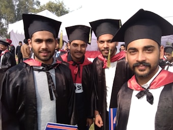Four young men wearing black graduation caps and gowns, with red and black collars, pose together outdoors during a graduation ceremony. In the background, more graduates and attendees can be seen standing near a white tent.