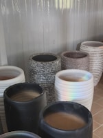 A collection of large, cylindrical pots in varied textures and colors, including black, white, speckled gray, and white with horizontal grooves. They are displayed against a corrugated metal backdrop, and the lighting casts a subtle rainbow reflection on one white pot.