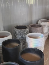 A collection of large, cylindrical pots in varied textures and colors, including black, white, speckled gray, and white with horizontal grooves. They are displayed against a corrugated metal backdrop, and the lighting casts a subtle rainbow reflection on one white pot.