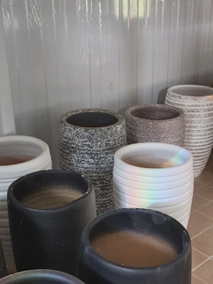 A collection of large, cylindrical pots in varied textures and colors, including black, white, speckled gray, and white with horizontal grooves. They are displayed against a corrugated metal backdrop, and the lighting casts a subtle rainbow reflection on one white pot.