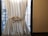 A white curtain tied with a bow partially covers a window, allowing some light to filter through. The outside view shows a metal staircase and urban building backdrop. The setting has a minimalistic and domestic feel, with the curtain framing the window strongly.