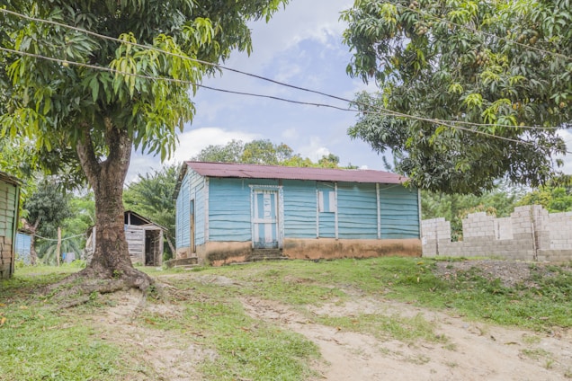 A small, simple house with light blue wooden siding and a rusty metal roof sits on a patch of land. Surrounding the house are green trees and a dirt path leading up to it. There is a partially constructed concrete block wall to the right and some smaller structures in the background.