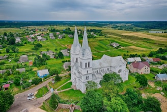 Aerial view of a large, white Gothic-style church with two tall spires, set amidst a rural landscape with lush green fields and small, scattered houses. The surrounding area shows a mix of grassy land and a few trees, with a road passing by the church.