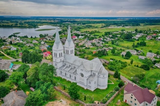A large, white, twin-spire church stands prominently amidst a verdant landscape. Surrounding the church are scattered houses and lush green fields, with a body of water visible in the distance. The sky is overcast, adding a serene and calm vibe to the rural scenery.