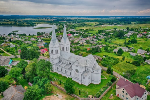 A large, white, twin-spire church stands prominently amidst a verdant landscape. Surrounding the church are scattered houses and lush green fields, with a body of water visible in the distance. The sky is overcast, adding a serene and calm vibe to the rural scenery.