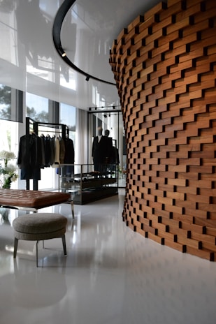 A modern interior space featuring a unique wooden wall with a brick-like pattern. The room includes a clothing display with garments on racks, a leather bench, and a round upholstered stool. The polished white floor and large windows create a bright and airy atmosphere with reflections of natural light.