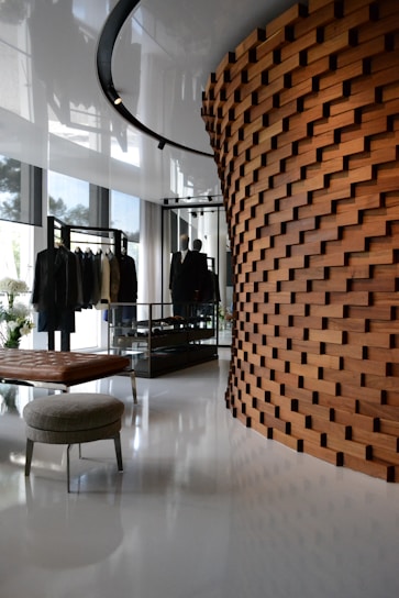 A modern interior space featuring a unique wooden wall with a brick-like pattern. The room includes a clothing display with garments on racks, a leather bench, and a round upholstered stool. The polished white floor and large windows create a bright and airy atmosphere with reflections of natural light.