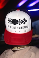 A person wearing a hat with a unique pattern and text design. The hat is mainly white with a red brim and features abstract black shapes along with the text 'U (KLIP) N U LEARN.' followed by numbers '11 12 9 22'. The person also wears gold chain necklaces.