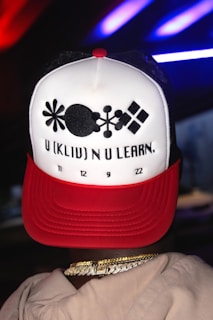 A person wearing a hat with a unique pattern and text design. The hat is mainly white with a red brim and features abstract black shapes along with the text 'U (KLIP) N U LEARN.' followed by numbers '11 12 9 22'. The person also wears gold chain necklaces.