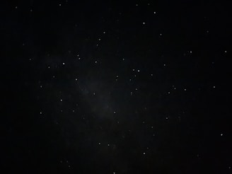 A dark night sky sprinkled with numerous small, bright stars. There are areas of varying darkness, suggesting the presence of distant nebulae.