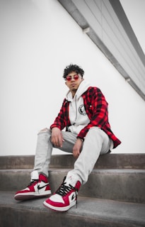 A person is sitting on concrete steps wearing stylish casual attire, including a red and black plaid jacket, a white hoodie, light gray ripped jeans, and red and white high-top sneakers. The individual is also sporting round red sunglasses and curly hair. The setting appears modern and minimalist with a clean, white background.