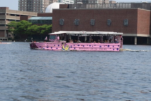 A pink amphibious tour vehicle, commonly known as a duck boat, is traveling on a body of water. It is filled with passengers and has 'Boston Duck Tours' written on its side. The background features a brick building with green trees and a modern cityscape.