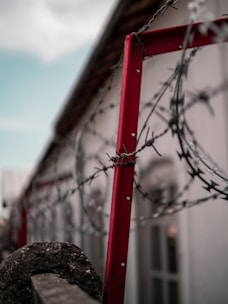 A close-up view of barbed wire coiled around a red metal frame, with a blurred building in the background. The image conveys a sense of confinement or security.