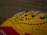 A close-up view of a yellow volleyball with visible lacing and printed text indicating 'Made in India' and 'Size 4'. The image includes some handwritten text and a portion of a red label on the ball.