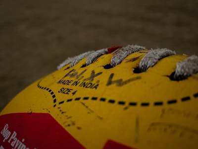 A close-up view of a yellow volleyball with visible lacing and printed text indicating 'Made in India' and 'Size 4'. The image includes some handwritten text and a portion of a red label on the ball.