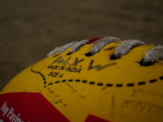 A close-up view of a yellow volleyball with visible lacing and printed text indicating 'Made in India' and 'Size 4'. The image includes some handwritten text and a portion of a red label on the ball.