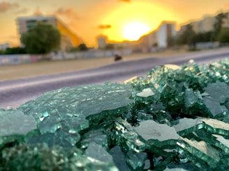 A close-up view of broken green glass shards in sharp focus, with an out-of-focus background showing a cityscape at sunset. The sun is low on the horizon, casting a warm golden glow over the scene. There are buildings in the distance with some greenery visible.