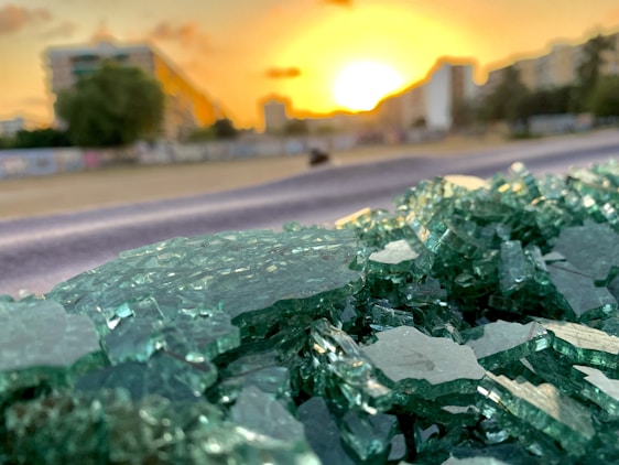 A close-up view of broken green glass shards in sharp focus, with an out-of-focus background showing a cityscape at sunset. The sun is low on the horizon, casting a warm golden glow over the scene. There are buildings in the distance with some greenery visible.