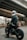 A person wearing a black leather jacket and a white helmet with an orange visor sits on a motorcycle under an overpass. The setting is urban, with concrete structures visible in the background. The motorcycle has a modern, rugged design with visible shock absorbers and a prominent rear tire. Another figure is seen walking in the distance.