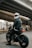 A person wearing a black leather jacket and a white helmet with an orange visor sits on a motorcycle under an overpass. The setting is urban, with concrete structures visible in the background. The motorcycle has a modern, rugged design with visible shock absorbers and a prominent rear tire. Another figure is seen walking in the distance.