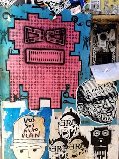 A vibrant street art mural featuring a large pink pixelated face with black eyes and mouth. Surrounding it are various smaller stencil art pieces including faces, text in Spanish, and abstract designs. The background is a mix of peeling paint and layered stickers with shades of blue and black.