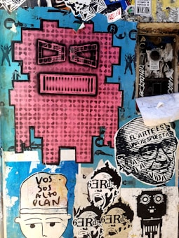 A vibrant street art mural featuring a large pink pixelated face with black eyes and mouth. Surrounding it are various smaller stencil art pieces including faces, text in Spanish, and abstract designs. The background is a mix of peeling paint and layered stickers with shades of blue and black.