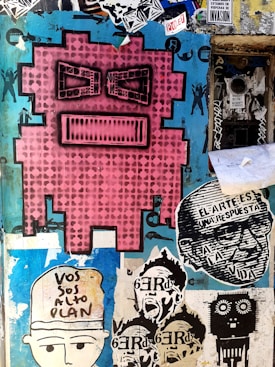 A vibrant street art mural featuring a large pink pixelated face with black eyes and mouth. Surrounding it are various smaller stencil art pieces including faces, text in Spanish, and abstract designs. The background is a mix of peeling paint and layered stickers with shades of blue and black.