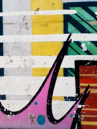 Colorful graffiti features a mix of geometric shapes with a large black swoosh and detailed weathering. Prominent colors include a mustard yellow block, a teal striped section, and patches of pink with blue dots. The paint is peeling, revealing layers underneath.