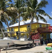 A bright yellow two-story building is surrounded by tall palm trees under a clear blue sky. In front of the building, there are two boats on trailers, and a sign that reads 'Fishing Supplies' is displayed next to vibrant pink flowers.