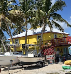 A bright yellow two-story building is surrounded by tall palm trees under a clear blue sky. In front of the building, there are two boats on trailers, and a sign that reads 'Fishing Supplies' is displayed next to vibrant pink flowers.