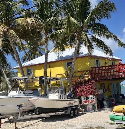 A bright yellow two-story building is surrounded by tall palm trees under a clear blue sky. In front of the building, there are two boats on trailers, and a sign that reads 'Fishing Supplies' is displayed next to vibrant pink flowers.
