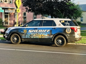 A sheriff's vehicle parked on the side of a street with a detailed decal depicting a cityscape and the text 'Sheriff Erie County' on the side. The vehicle is a white and blue SUV with official badges and decals, parked near a pink building with a coffee house sign. The environment is a suburban area with trees and street signs visible.