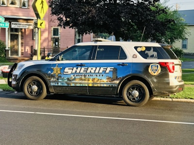 A sheriff's vehicle parked on the side of a street with a detailed decal depicting a cityscape and the text 'Sheriff Erie County' on the side. The vehicle is a white and blue SUV with official badges and decals, parked near a pink building with a coffee house sign. The environment is a suburban area with trees and street signs visible.