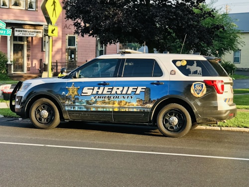 A sheriff's vehicle parked on the side of a street with a detailed decal depicting a cityscape and the text 'Sheriff Erie County' on the side. The vehicle is a white and blue SUV with official badges and decals, parked near a pink building with a coffee house sign. The environment is a suburban area with trees and street signs visible.