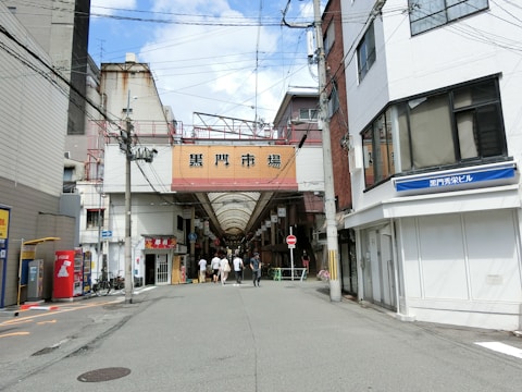 An urban street scene featuring a covered market entrance beneath a large sign. Buildings line both sides of the street, with various signs and advertisements visible. Several people walk towards the market in the background, and vending machines are seen on the left side.