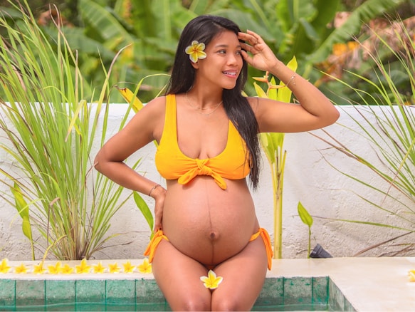A pregnant woman wearing an orange bikini sits by a pool adorned with yellow plumeria flowers. She has a flower tucked behind her ear and is gently touching her head while smiling. The background features lush green plants and a white wall.