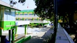 A modern, three-story building with a green and white color scheme is situated within a campus or school environment. The architecture features clean lines and a shaded walkway with trees and a flagpole in the courtyard. Motorcycles are parked along a pathway, indicating it might be a busy area.